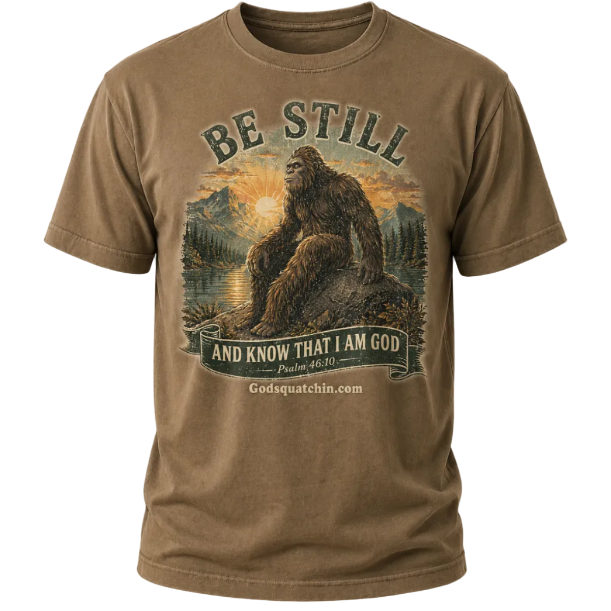 Brown t-shirt featuring a vintage-style graphic of Bigfoot sitting calmly on a rock beside a mountain lake at sunrise, surrounded by pine trees and mountains, with the text “Be Still and Know That I Am God – Psalm 46:10”