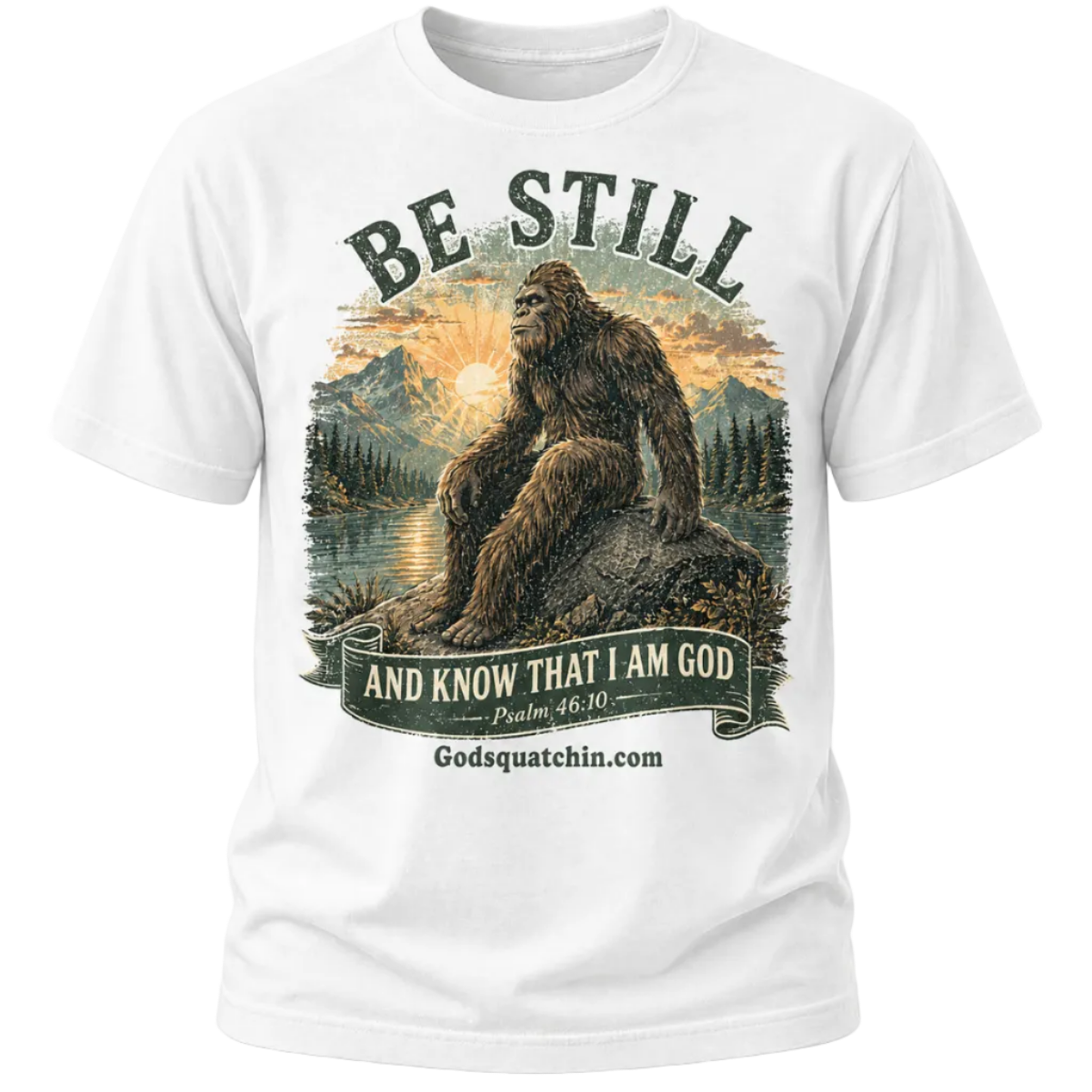 White t-shirt featuring a vintage-style graphic of Bigfoot sitting calmly on a rock beside a mountain lake at sunrise, surrounded by pine trees and mountains, with the text “Be Still and Know That I Am God – Psalm 46:10”